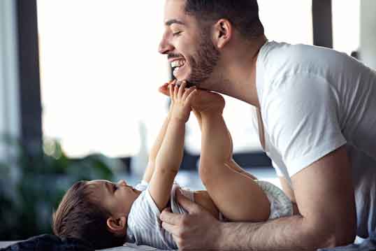 A joyful moment between a father and baby, both enjoying playful time in nappies on a bed.